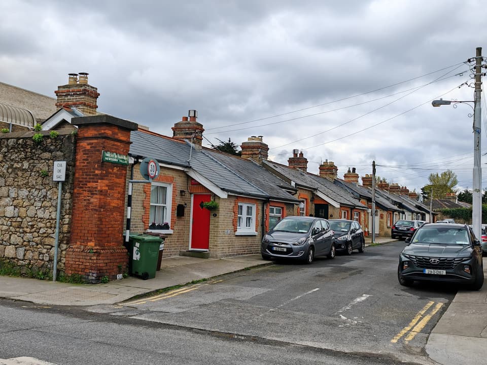 A row of single-storey red-brick cottages with parked cars on a street in Blackrock, Dublin.