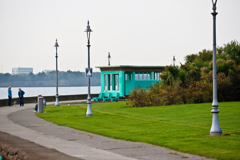 The seafront promenade at Clontarf with a lawn, lamp posts and a turquoise shelter beside Dublin Bay.