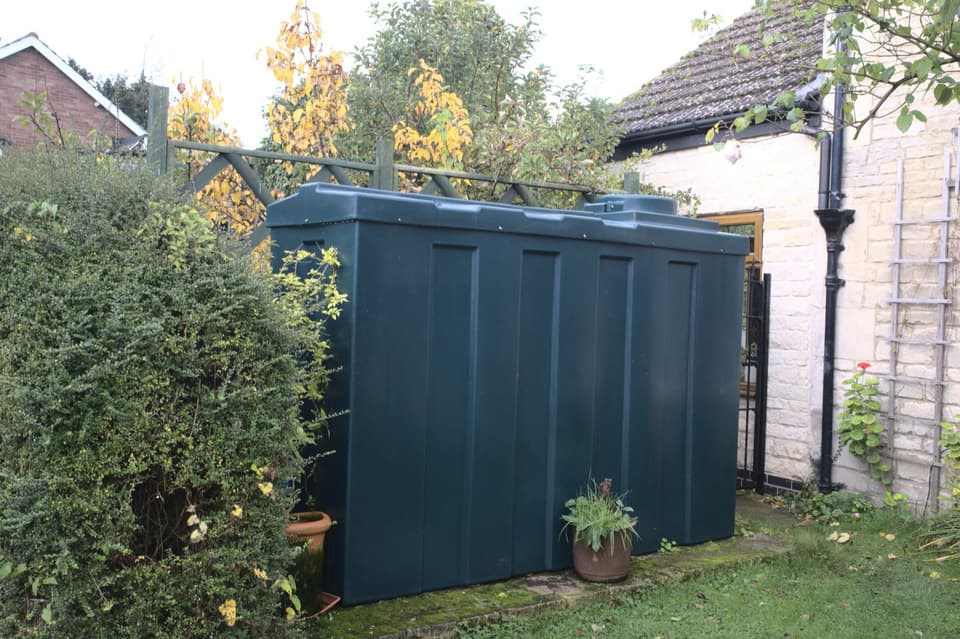 A dark green plastic heating oil tank standing on a slab in a garden beside a house wall.