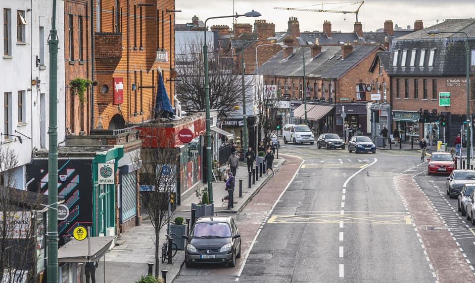 Ranelagh's main street in Dublin with red-brick shops, parked cars and a few pedestrians.