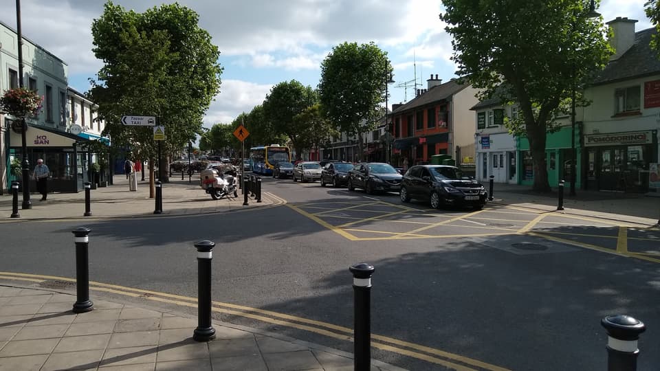 Swords Main Street at a junction, with shops, trees and parked cars on a sunny day.